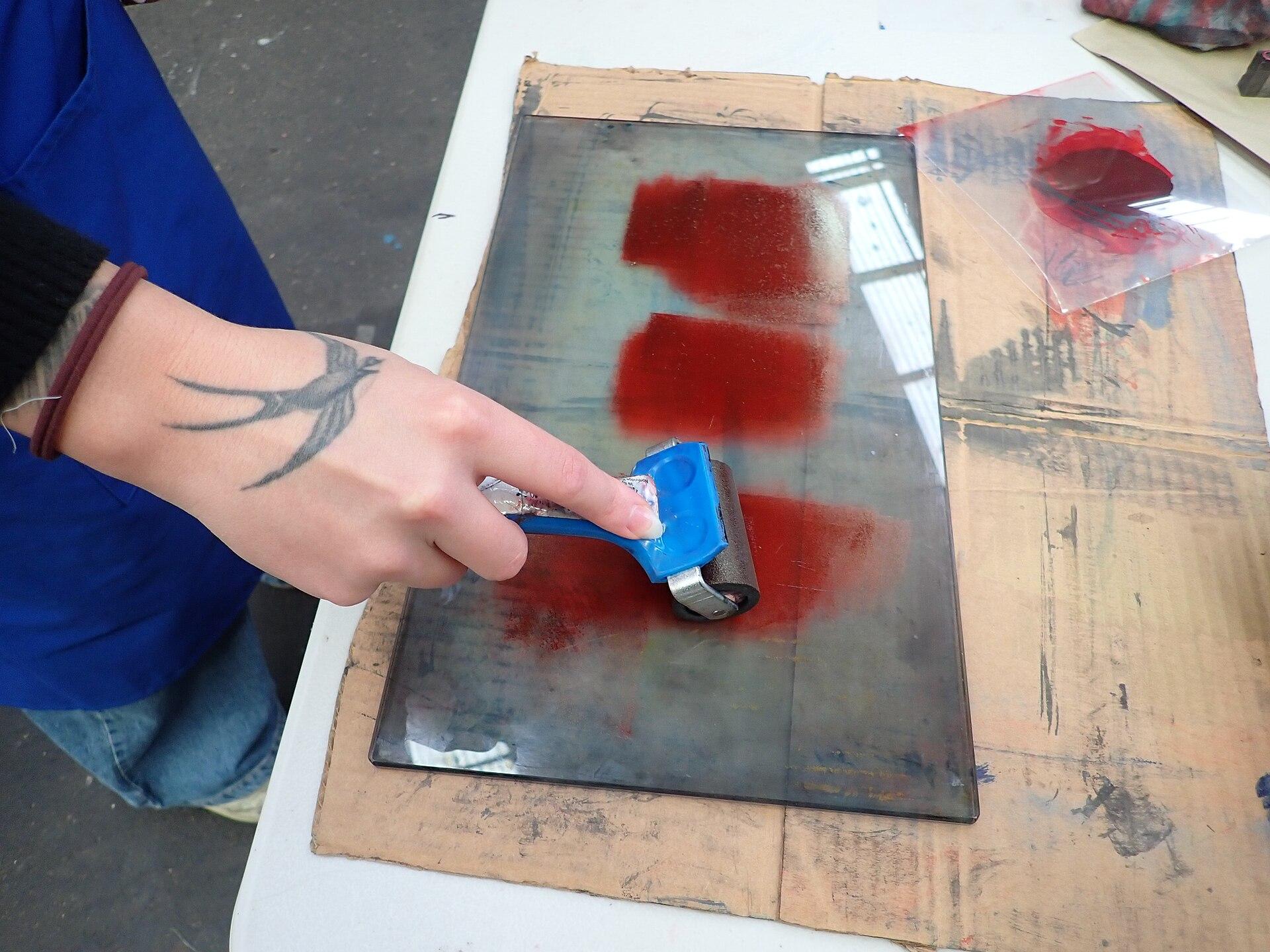 A hand rolling red ink out on a glass slab with a blue brayer